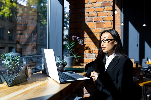 Biznes kobieta freelancer pracująca zdalnie w kawiarni na laptopie, zamyślona i skupiona Azjatka rozmawiająca podczas wideorozmowy z kolegami, patrząca na kamerę w laptopie