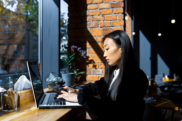 Biznes kobieta freelancer, pracująca zdalnie w kawiarni na laptopie, zamyślona i skupiona Azjatka