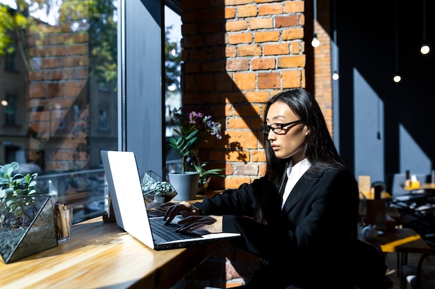 Biznes kobieta freelancer, pracująca zdalnie w kawiarni na laptopie, zamyślona i skupiona Azjatka