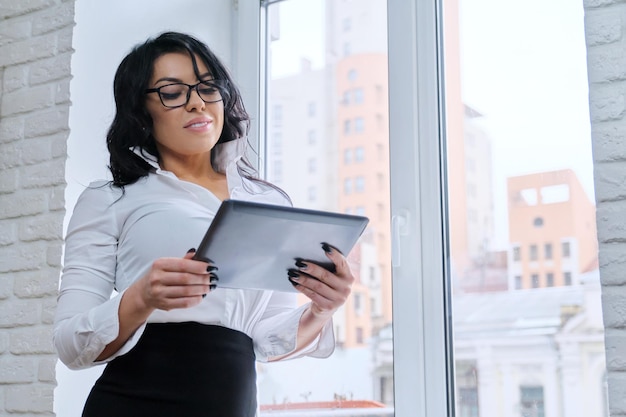 Biznes piękna kobieta w białej bluzce okulary z cyfrowego tabletu w biurze w pobliżu okna. Sukces, kariera, personel, profesjonalna kobieta