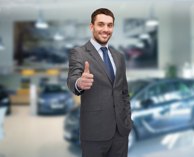 biznes samochodowy, sprzedaż samochodów, koncepcja gestów i ludzi - uśmiechnięty biznesmen pokazujący kciuki w górę na tle pokazu samochodowego