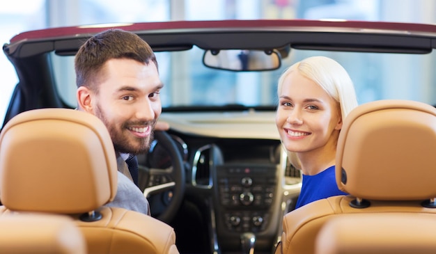 biznes samochodowy, sprzedaż samochodów, konsumpcjonizm i koncepcja ludzi - szczęśliwa para siedzi w samochodzie na pokazie samochodowym lub salonie