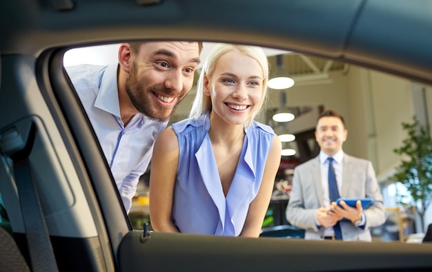 biznes samochodowy, sprzedaż samochodów, technologia i koncepcja ludzi - szczęśliwa para z dealerem samochodowym w salonie samochodowym lub salonie