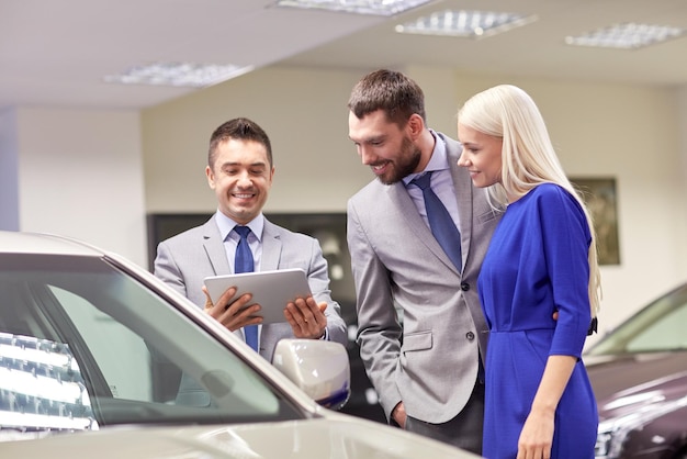 biznes samochodowy, sprzedaż samochodów, technologia i koncepcja ludzi - szczęśliwa para z dealerem samochodowym w salonie samochodowym lub salonie