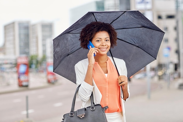 biznes, zła pogoda i ludzie i koncepcja - szczęśliwa afroamerykańska bizneswoman z parasolem i torebką dzwoniąca na smartfona na ulicy miasta