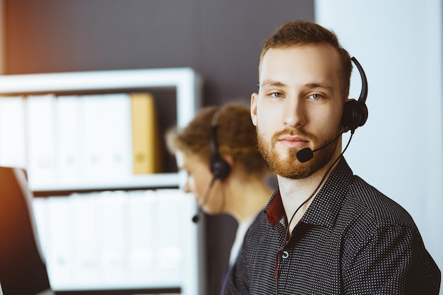 Biznesmen czerwonobrody rozmawia przez zestaw słuchawkowy w pobliżu koleżanki, siedząc w nowoczesnym biurze słonecznym. Zróżnicowana grupa ludzi w call center.