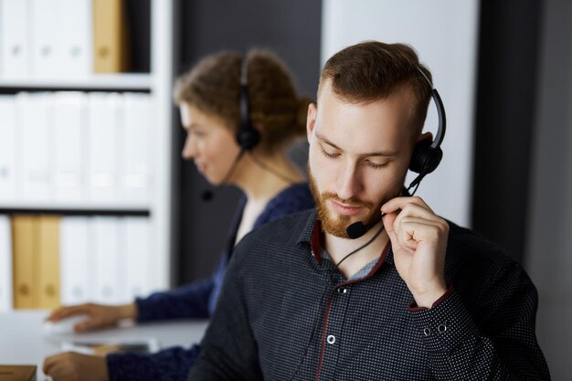 Biznesmen czerwonobrody rozmawia przez zestaw słuchawkowy w pobliżu koleżanki siedząc w nowoczesnym biurze. Zróżnicowana grupa ludzi w call center. Telemarketing i obsługa klienta.