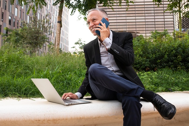 Biznesmen kaukaski za pomocą laptopa i rozmawia przez telefon komórkowy, siedząc na ławce w parku obok budynków biurowych. Koncepcja biznesu i technologii.
