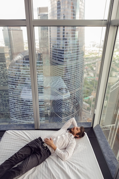 Biznesmen mężczyzna w białej koszuli leży na łóżku w pobliżu dużego panoramicznego okna odpoczywając