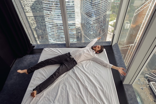 Biznesmen mężczyzna w białej koszuli leży na łóżku w pobliżu dużego panoramicznego okna odpoczywając