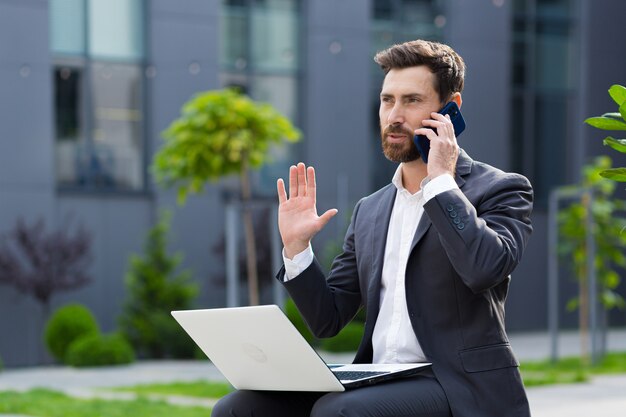 Biznesmen Odnoszący Sukcesy Zgłasza Dobre Wieści Przez Telefon, Pracując Z Laptopem W Porze Lunchu W Pobliżu Biura, Siedząc Na ławce