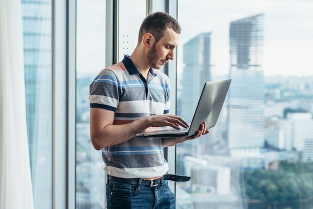 Biznesmen posiadający laptopa pracy stojącej w biurze w pobliżu okna z widokiem na miasto.