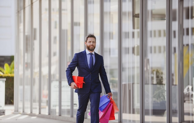 Biznesmen robi zakupy w centrum handlowym Szczęśliwy biznesmen w garniturze trzymający papierowe torebki Zakupoholik idący ulicą handlową z mnóstwem toreb na zakupy Bogacz