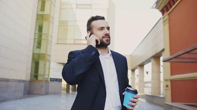 Biznesmen rozmawia przez telefon komórkowy, robiąc transakcje i spacerując w pobliżu nowoczesnych biurowców