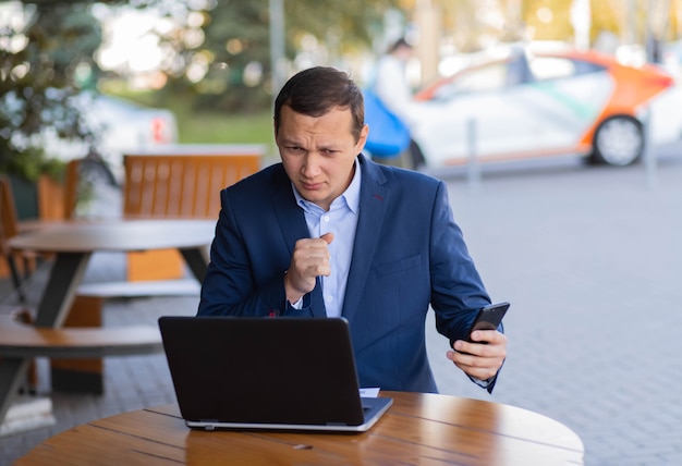 Biznesmen siedzi przy stoliku w kawiarni na ulicy, kaszle w dłoń, podczas przerwy w finansowej dzielnicy prowadzi rozmowę wideo przez telefon komórkowy. Choroba podczas pracy.