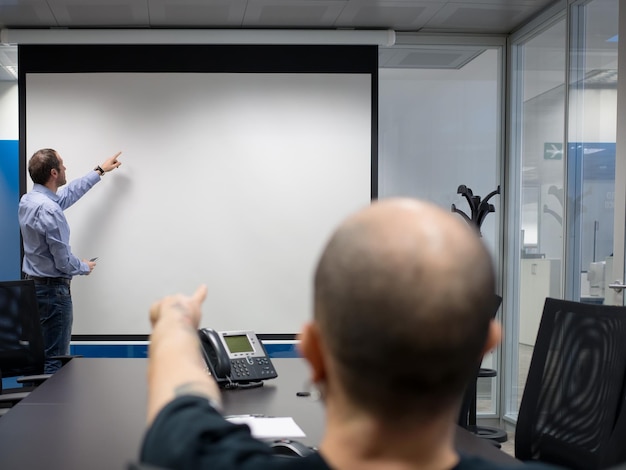 Biznesmen Stojący Przed Ekranem Projekcyjnym, Podczas Gdy Współpracownik Siedzi Przy Stole W Biurze