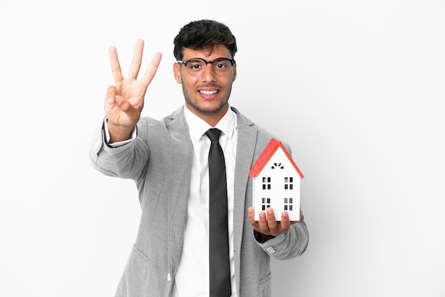 Biznesmen trzymający dom na białym tle na niebieskim tle szczęśliwy i liczący trzy palcami
