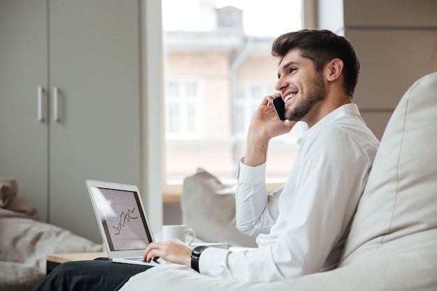 Biznesmen ubrany w białą koszulę siedzi w kawiarni rozmawia przez telefon podczas korzystania z laptopa.