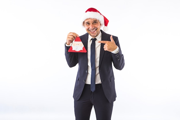 Biznesmen ubrany w garnitur, krawat i świąteczny kapelusz Świętego Mikołaja, uśmiechając się, wskazując na ozdobę Santa, na białym tle. Koncepcja obchody Bożego Narodzenia.