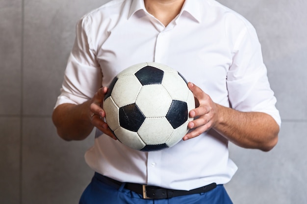 Biznesmen w białej koszuli stoi pod ścianą i trzyma w rękach piłkę nożną.