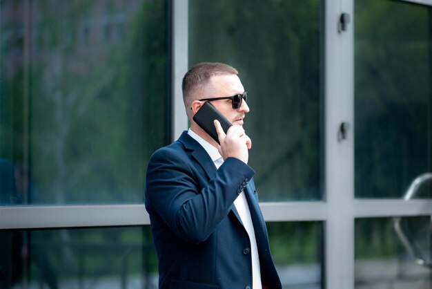 Biznesmen w ciemnych okularach przeciwsłonecznych z telefonem w ręku stoi przed budynkiem biznesowym i rozmawia przez telefon. Ubrany jest w lekką koszulę i marynarkę w stylu casual.