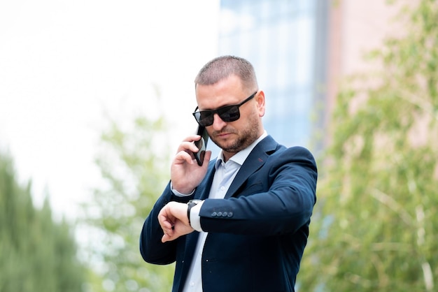 Biznesmen w ciemnych okularach stoi przed budynkiem firmy rozmawia przez telefon.
