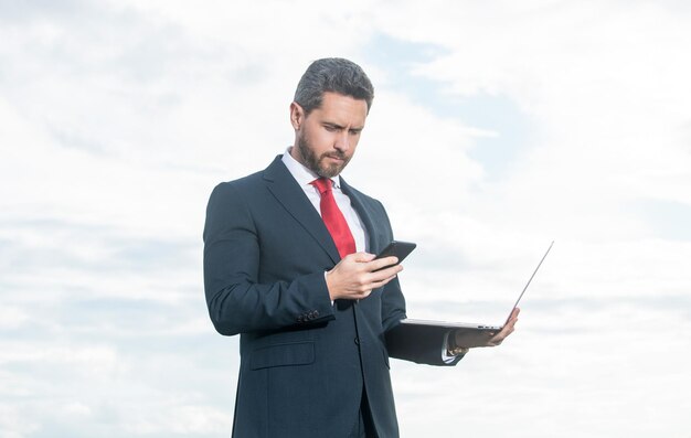 Biznesmen w garniturze sprawdza smartfona i trzyma laptopa