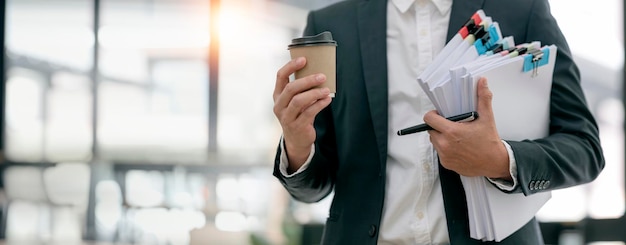 Biznesmen w garniturze trzymający duży stos dokumentów i filiżankę kawy stojący w biurze z miejscem na kopię Egzaminowanie i zarządzanie dokumentami