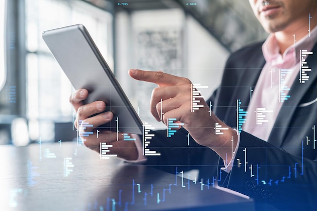 Biznesmen w garniturze za pomocą tabletu, aby zoptymalizować strategię handlową w hologramie wykresu Forex w funduszu finansów korporacyjnych na nowoczesnym, panoramicznym tle biurowym