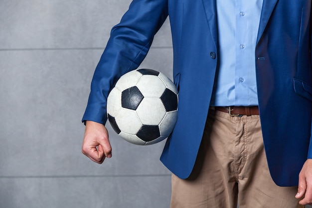 Biznesmen w niebieskiej marynarce stoi pod ścianą i trzyma w ręku piłkę nożną.