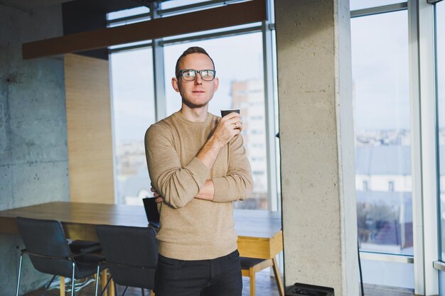 Biznesmen w okularach i stylowych ubraniach pije kawę w nowoczesnym miejscu do pracy Mężczyzna freelancer z laptopem pracuje zdalnie