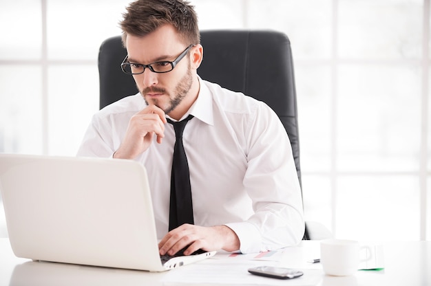 Biznesmen w pracy. Przystojny młody broda mężczyzna w koszuli i krawacie pracuje na laptopie, siedząc w swoim miejscu pracy