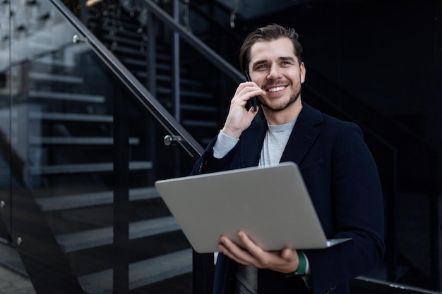 Biznesmen w średnim wieku rozmawia przez telefon komórkowy z szerokim uśmiechem i trzyma w rękach laptopa