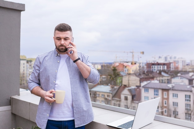 Biznesmen w ubranie relaksując się na tarasie z widokiem na miasto korzysta z telefonu i laptopa podczas picia kawy