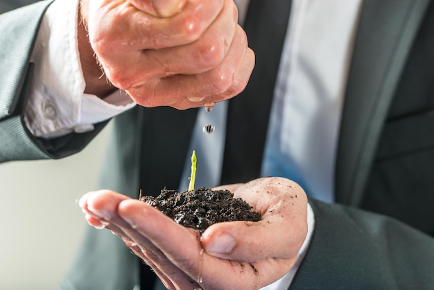 Biznesmen wlewający wodę nad małą zieloną rośliną rosnącą w żyznej glebie trzymanej w złożonej lewej ręce koncepcji ekologii środowiska i ekologicznego zbliżenie biznesowego