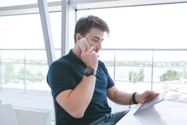 Biznesmen z brodą i zwykłą odzieżą siedzi wokół lekkiej kawiarni, patrzy na tablet i rozmawia przez telefon