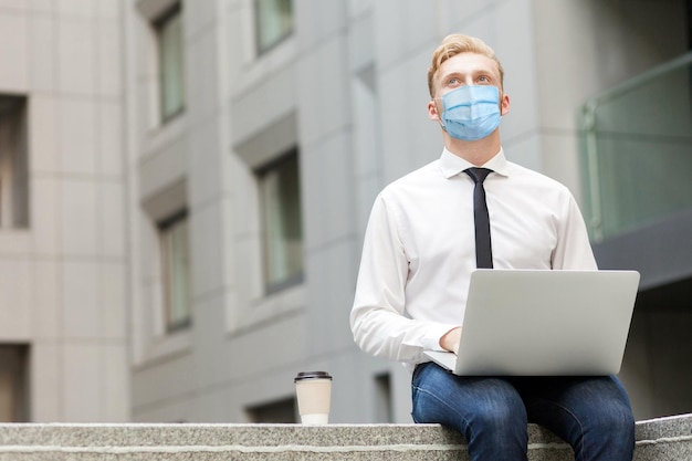 Biznesmen z chirurgiczną maską medyczną pracuje nad outsourcingiem laptopa i zastanawia się