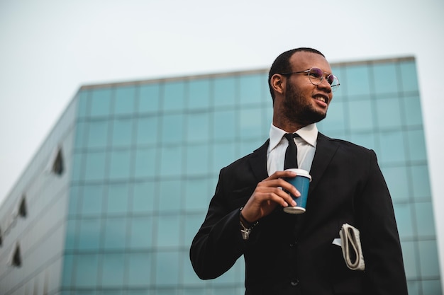 Zdjęcie biznesmen z kawą na wynos przed wieżowcem. czarny etniczny mężczyzna w marynarce i krawacie