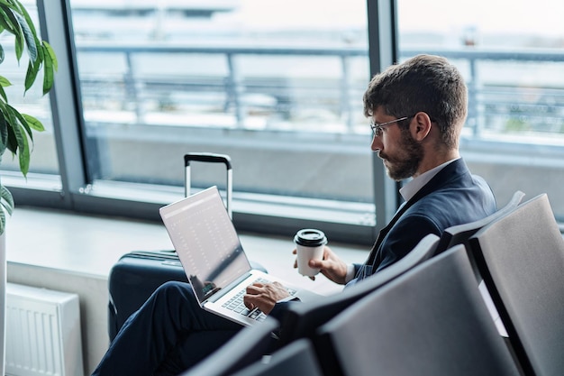 Biznesmen z widokiem z boku z kawą na wynos przy laptopie w hali odlotów na lotnisku