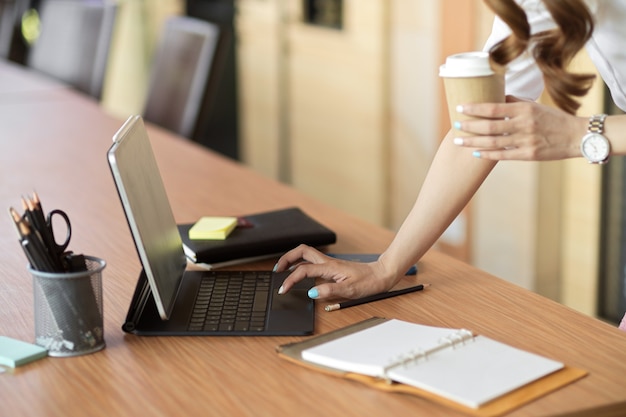 biznesowa kobieta opiera się na biurku, pisząc na klawiaturze tabletu, trzymając kawę na wynos