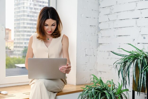 Biznesowa kobieta siedzi na parapecie w swoim biurze z laptopem na kolanach