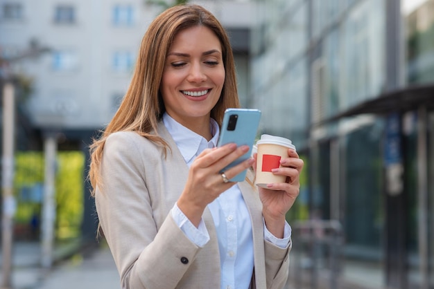 Biznesowa kobieta używająca telefonu do wysyłania wiadomości i trzymająca filiżankę kawy