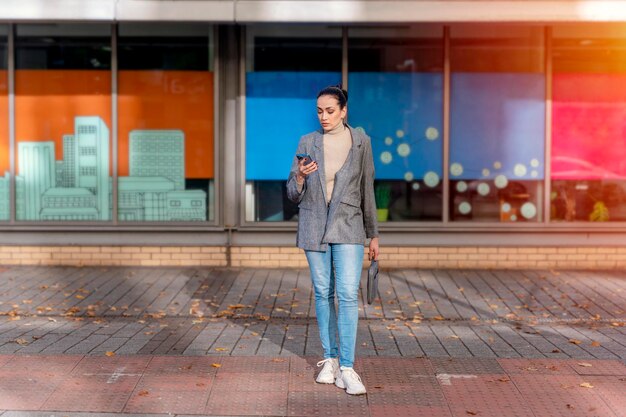 biznesowa kobieta w kurtce i dżinsach czeka na zielone światło drogowe, aby przejść przez ulicę i używać telefonu