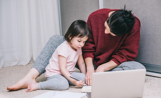 Biznesowa mama freelancer i jej mała ciekawska córka pracują razem na podłodze w domu i używają laptopa i rysują coś na papierze Kobieta interesu pracuje w domu z dzieckiem