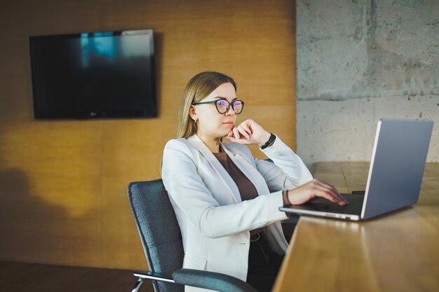 Biznesowa młoda kobieta w okularach i przypadkowych ubraniach siedzi przy drewnianym stole z laptopem i filiżanką kawy odrabianiu lekcji na laptopie