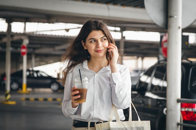 Biznesowa młoda kobieta z kawą rozmawia radośnie przez telefon na parkingu