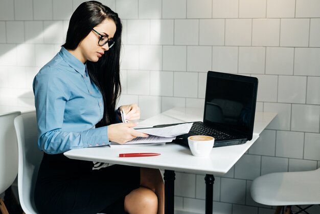 Biznesowa młoda studentka w niebieskiej koszuli i okularach pracuje na laptopie w kawiarni
