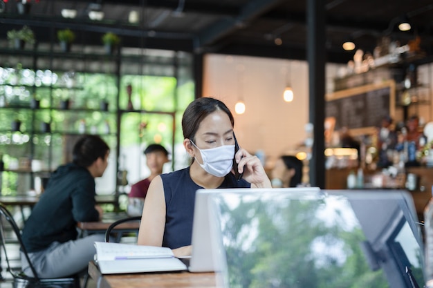 Bizneswoman jest ubranym twarzy maskę opowiada na telefonie komórkowym pracuje na laptopie.