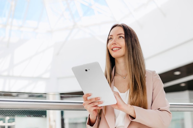 Bizneswoman stoi w pomieszczeniu w budynku biurowym, trzymając tabletkę i uśmiechając się