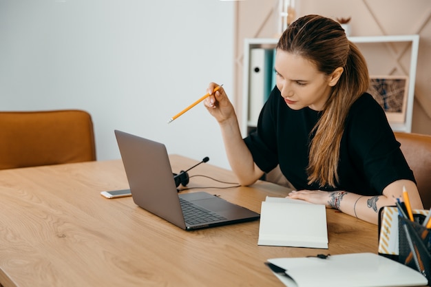 Bizneswoman W Słuchawkach Siedzi Przy Biurku, Patrzy Na Ekran Laptopa, Robiąc Notatki